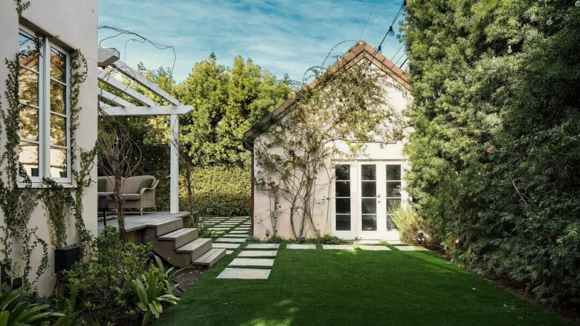 Backyard lawn with a detached white stucco structure with French doors, a pergola and a hedge behind it