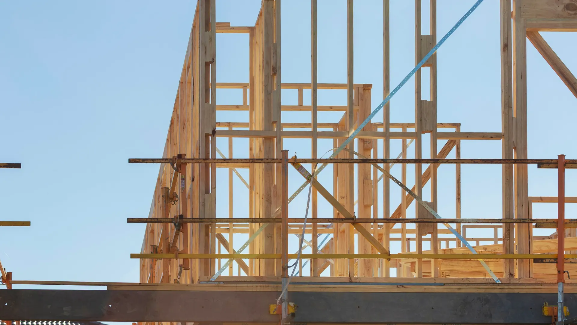 Steel scaffold rails in front of wood-framed upper-floor walls with metal strap bracing, under a clear blue sky